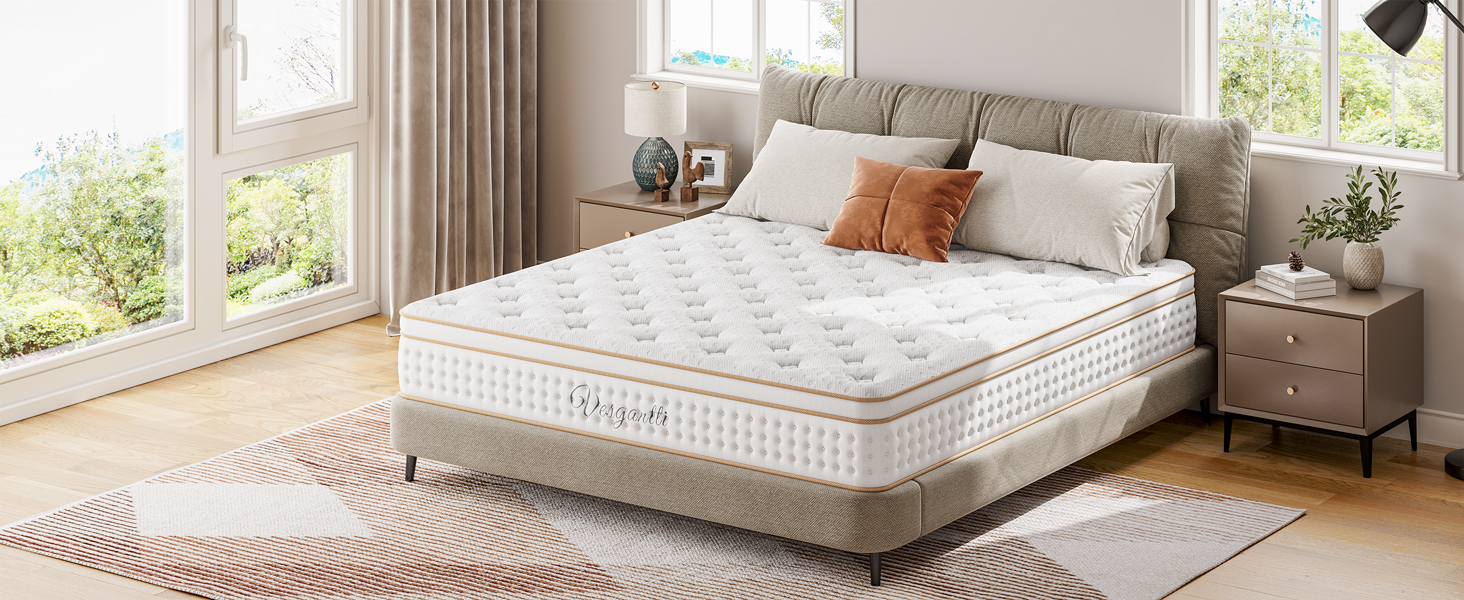 Plush mattress with quilted top pattern on grey upholstered bed frame, styled with beige bedding and orange accent pillow in bright bedroom setting.